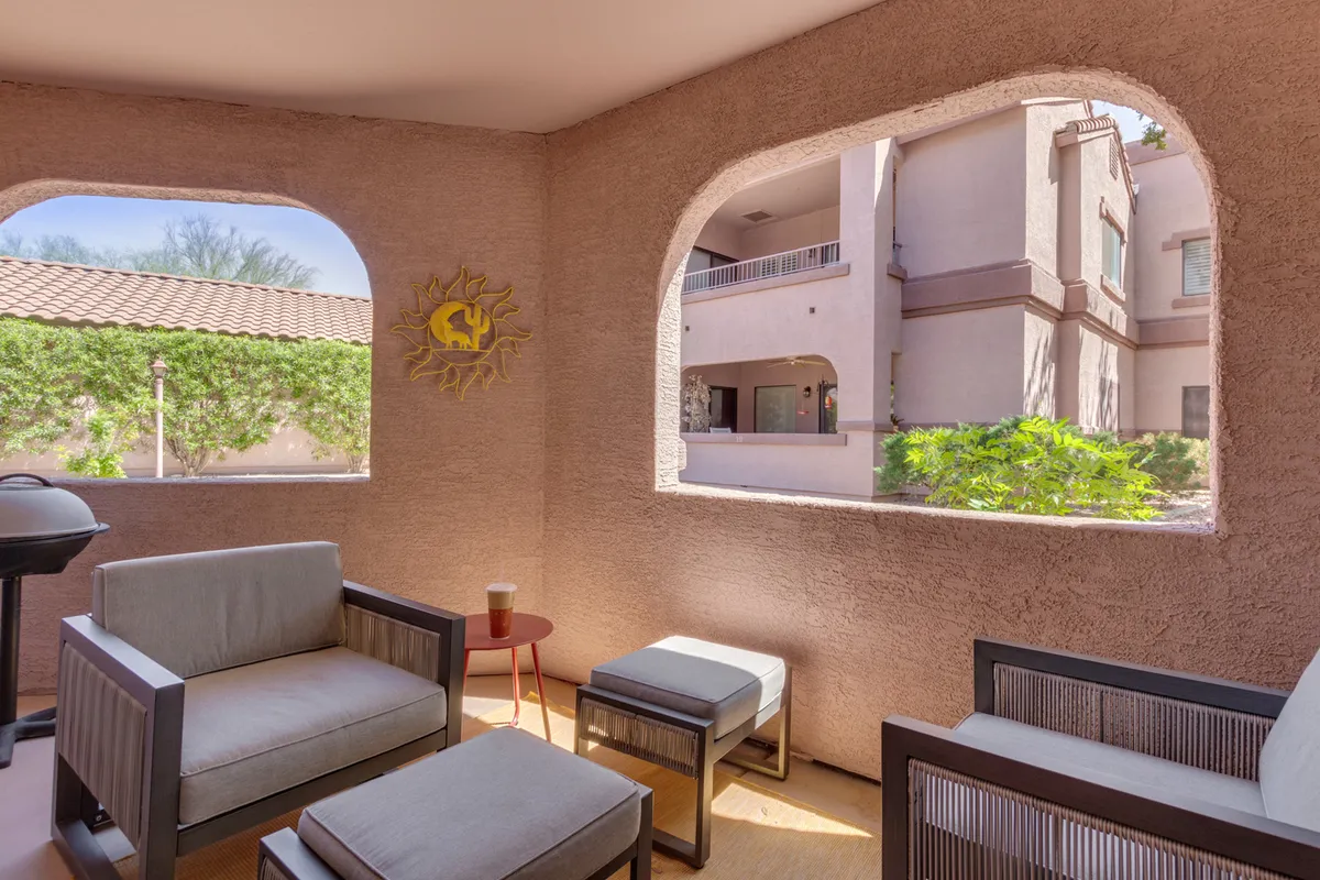 A bright, sunlit area with arched windows reveals comfortable outdoor seating and a decorative wall sun. This space bridges the indoor and outdoor experience, suggesting a spot for morning coffee or evening relaxation while enjoying views of the community and sky.