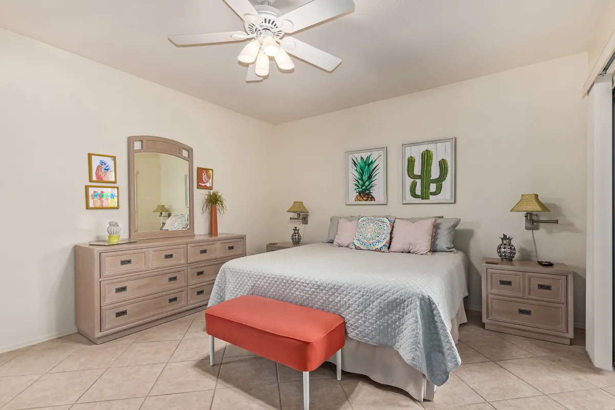 A cozy, inviting bedroom featuring a plush bed with soft bedding, a dresser and mirror, accent artwork, and a cushioned bench at the foot of the bed. Natural light gently fills the space, creating a restful ambiance ideal for relaxation or winding down with a book.
