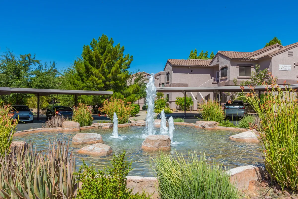 An inviting exterior view of a landscaped community garden complete with a centerpiece fountain, surrounding greenery, and visible community buildings. The space is peaceful and communal, suggesting opportunities for families to stroll, relax, and interact in a secure setting.