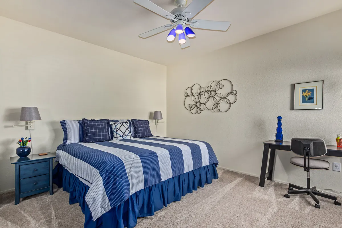 A peaceful bedroom with a bed adorned in blue striped bedding, complemented by a blue nightstand with a flower vase and a small desk with decorative accents. The overall setting is neat and calming, ideal for restful nights or productive mornings.